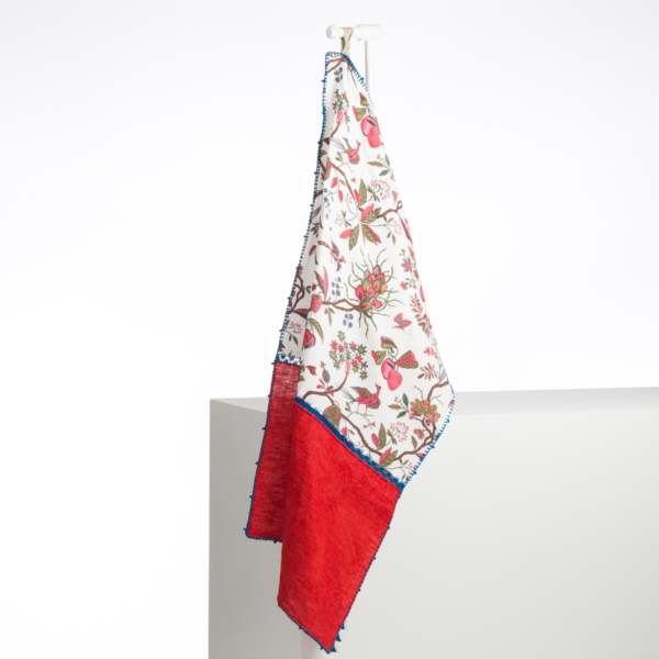 L'ABAT-JOUR SUR COMMANDE est un textile décoratif aux motifs floraux et d'oiseaux sur fond blanc, rouge en bas et bleu sur les bords. Il est suspendu à un crochet blanc sur un fond blanc uni. la marque Toile de Jouy