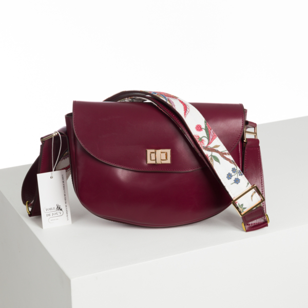 Le sac ABAT-JOUR SUR COMMANDE est un sac à bandoulière en cuir rouge foncé, doté d'un fermoir doré et d'une large lanière blanche ornée d'un motif floral coloré. Il est accompagné d'une étiquette et présenté sur une surface blanche. la marque Toile de Jouy