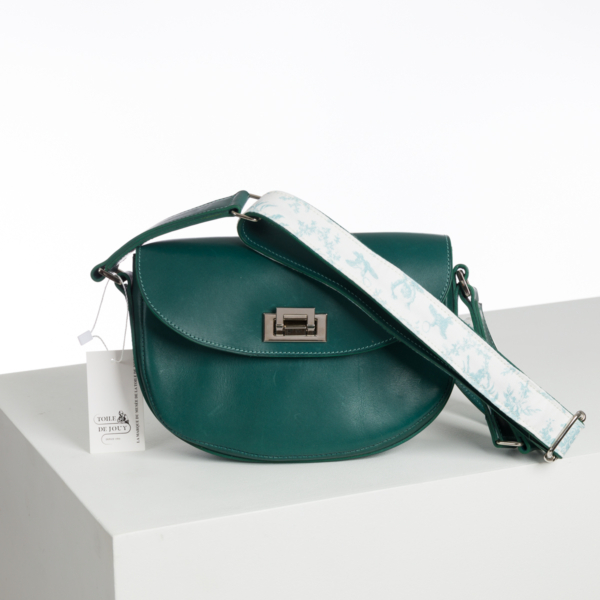 Un sac à bandoulière en cuir sarcelle nommé ABAT-JOUR SUR COMMANDE est doté d'un fermoir argenté et d'une large lanière blanche avec un motif bleu clair. Il repose sur une surface blanche avec une étiquette indiquant "L'OEIL DE DIOGENE". la marque Toile de Jouy