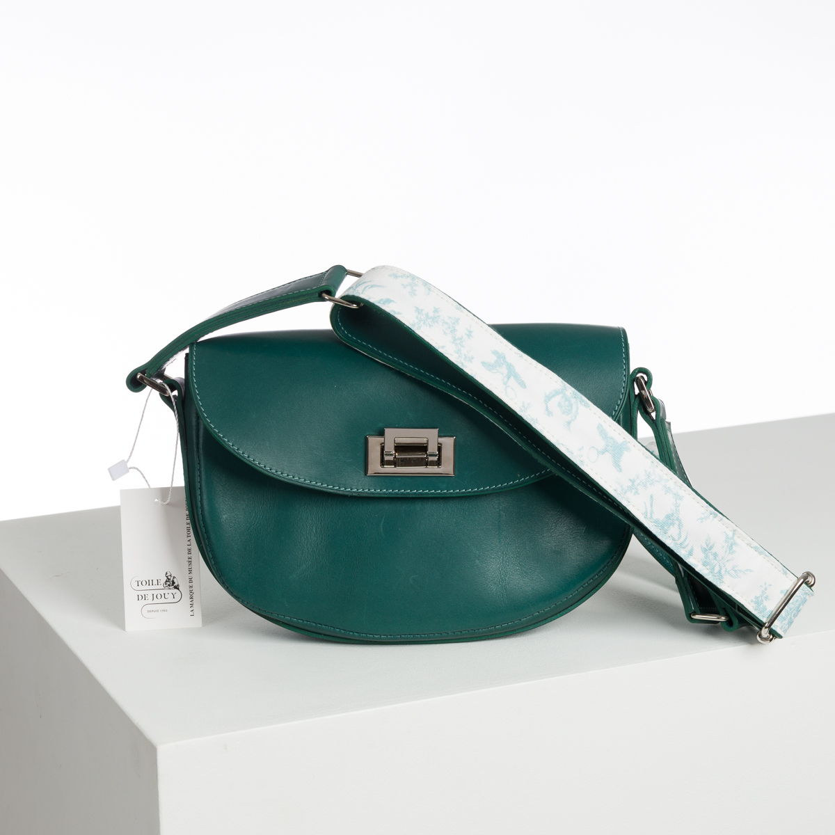 Un sac à bandoulière en cuir sarcelle nommé ABAT-JOUR SUR COMMANDE est doté d'un fermoir argenté et d'une large lanière blanche avec un motif bleu clair. Il repose sur une surface blanche avec une étiquette indiquant "L'OEIL DE DIOGENE". la marque Toile de Jouy