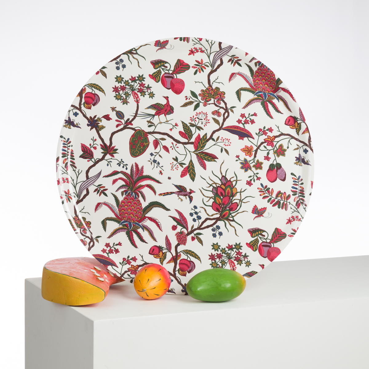 L'assiette ronde ABAT-JOUR SUR COMMANDE, au motif floral et fruitier joyeux, se tient debout sur une surface blanche, accompagnée de trois fruits en bois peints plus vrais que nature et d'un morceau de fromage réaliste. la marque Toile de Jouy