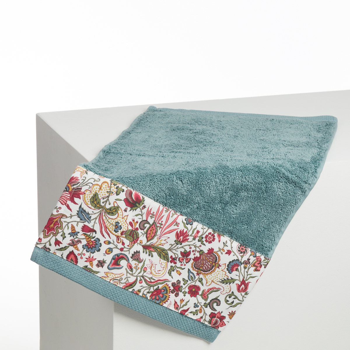L'ABAT-JOUR SUR COMMANDE est représenté drapé sur le bord d'une surface blanche, avec un tissu sarcelle et une bordure à motifs floraux rouges, roses, verts et jaunes sur un fond uni. la marque Toile de Jouy