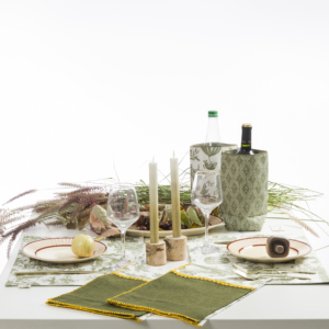 Une table à manger soigneusement dressée avec des accents verts, des verres à vin, des bougies dans des supports en bois, une bouteille de vin dans un manchon à motifs, deux serviettes de table vertes, une assiette de pain et des compositions d'herbes décoratives. la marque Toile de Jouy