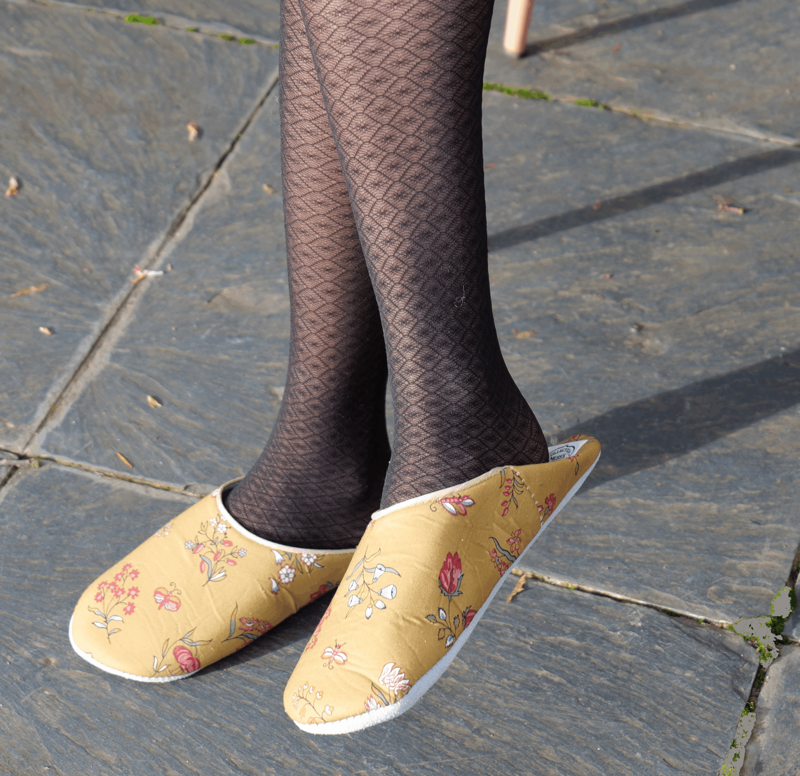 Une personne portant des collants noirs à motifs et des pantoufles BABOUCHES "SEMIS FLORAL" jaunes à fleurs se tient sur un trottoir en pierre, les jambes croisées à la cheville. la marque Toile de Jouy