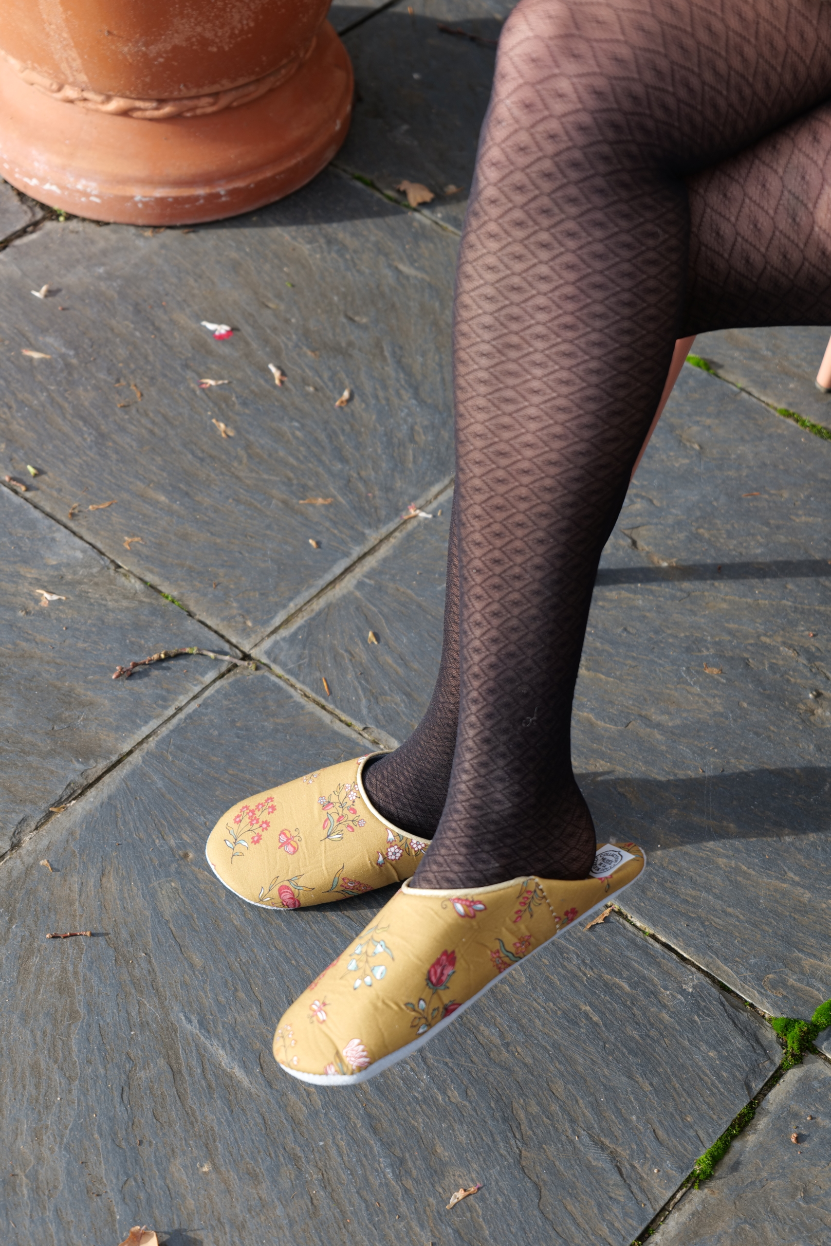 Une personne portant des collants noirs à motifs et des pantoufles à fleurs jaunes BABOUCHES "SEMIS FLORAL" est assise les jambes croisées sur un patio en pierre, une partie d'un grand pot en terre cuite étant visible dans le coin supérieur gauche. la marque Toile de Jouy