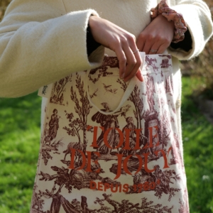 Une personne vêtue d'un manteau de couleur crème se tient debout sur une pelouse verte, avec à la main le TOTE BAG "TOILE DE JOUY DEPUIS 1760", qui comporte un motif et un texte imprimé en rouge. la marque Toile de Jouy