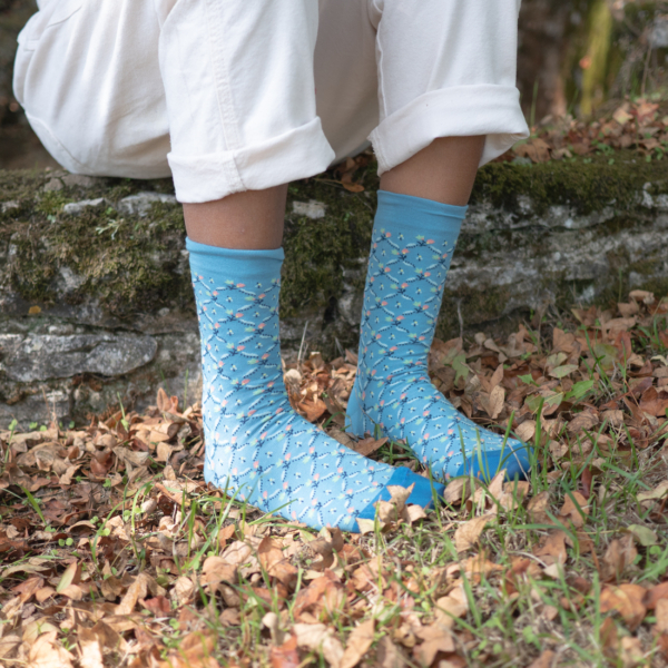 Personne assise en plein air sur un rebord de pierre, vêtue d'un pantalon blanc retroussé et de chaussettes bleues avec un petit motif floral. Des feuilles sèches et de l'herbe couvrent le sol autour de ses pieds. la marque Toile de Jouy