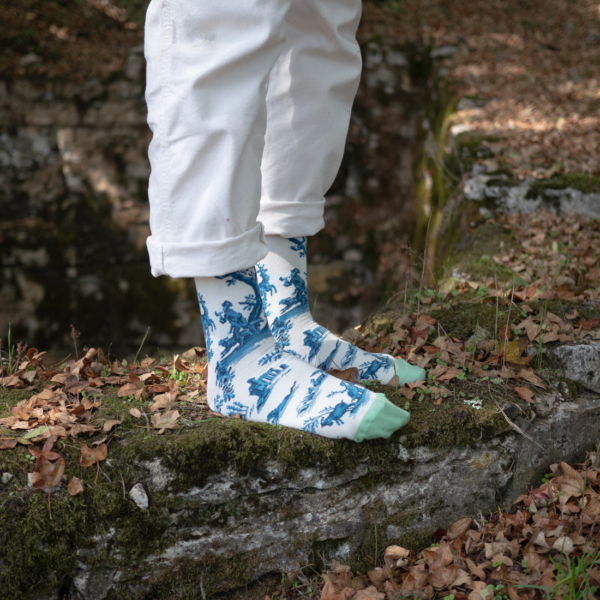 Une personne portant un pantalon blanc et des chaussettes à motifs bleus et blancs avec des orteils verts se tient sur une pierre moussue entourée de feuilles brunes tombées à l'extérieur. la marque Toile de Jouy