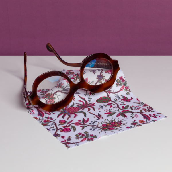La XCOMM_MICROFIBRE ANANAS, une paire de lunettes rondes de couleur marron, est présentée sur un tissu blanc avec un motif floral vibrant sur une surface blanche et sur un fond violet. la marque Toile de Jouy