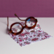 La XCOMM_MICROFIBRE ANANAS, une paire de lunettes rondes de couleur marron, est présentée sur un tissu blanc avec un motif floral vibrant sur une surface blanche et sur un fond violet. la marque Toile de Jouy