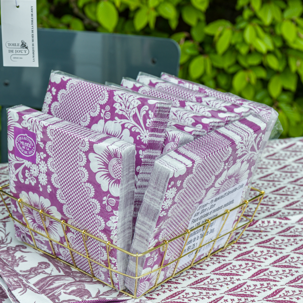 Un panier XCOMM_MICROFIBRE ANANAS contient des piles de serviettes en papier à motifs floraux violets et blancs sur une nappe assortie, avec du feuillage vert en arrière-plan. la marque Toile de Jouy