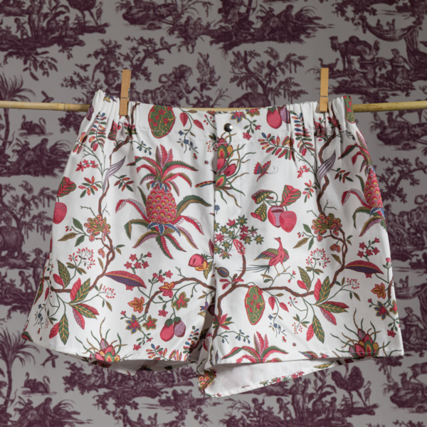 Un boxer blanc XCOMM_MICROFIBRE ANANAS avec un imprimé floral et de fruits tropicaux est suspendu à une corde à linge contre un mur à motifs de toile vintage. la marque Toile de Jouy