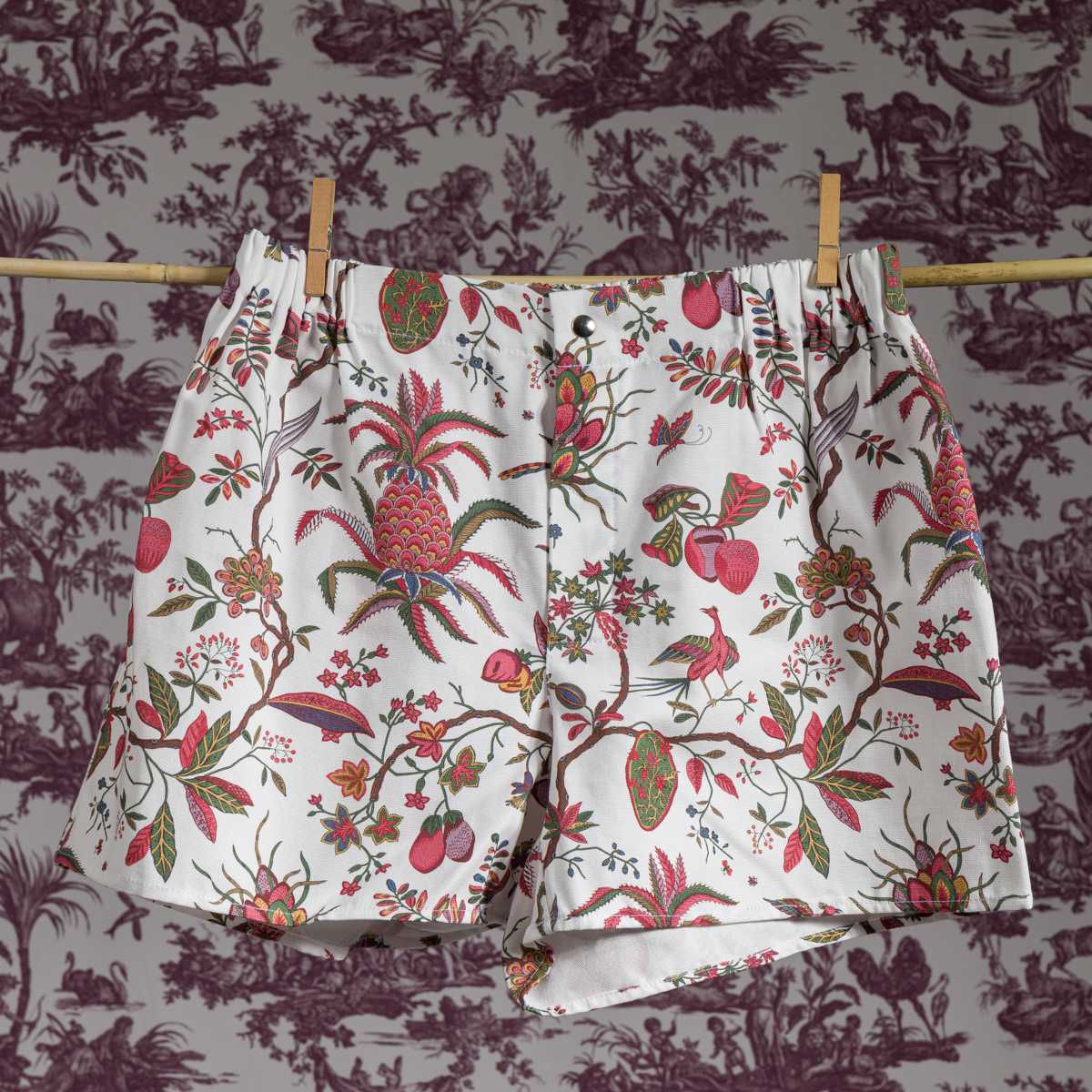 Un boxer blanc XCOMM_MICROFIBRE ANANAS avec un imprimé floral et de fruits tropicaux est suspendu à une corde à linge contre un mur à motifs de toile vintage. la marque Toile de Jouy
