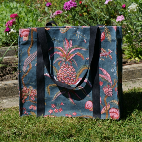 Un sac XCOMM_SAC CABAS MEDIUM ANANAS-FLEURS STYLISÉES - TISSAGE, un sac fourre-tout en tissu de taille moyenne orné d'un motif stylisé d'ananas et de fleurs roses et vertes, est posé sur l'herbe devant un parterre agrémenté de fleurs roses. la marque Toile de Jouy