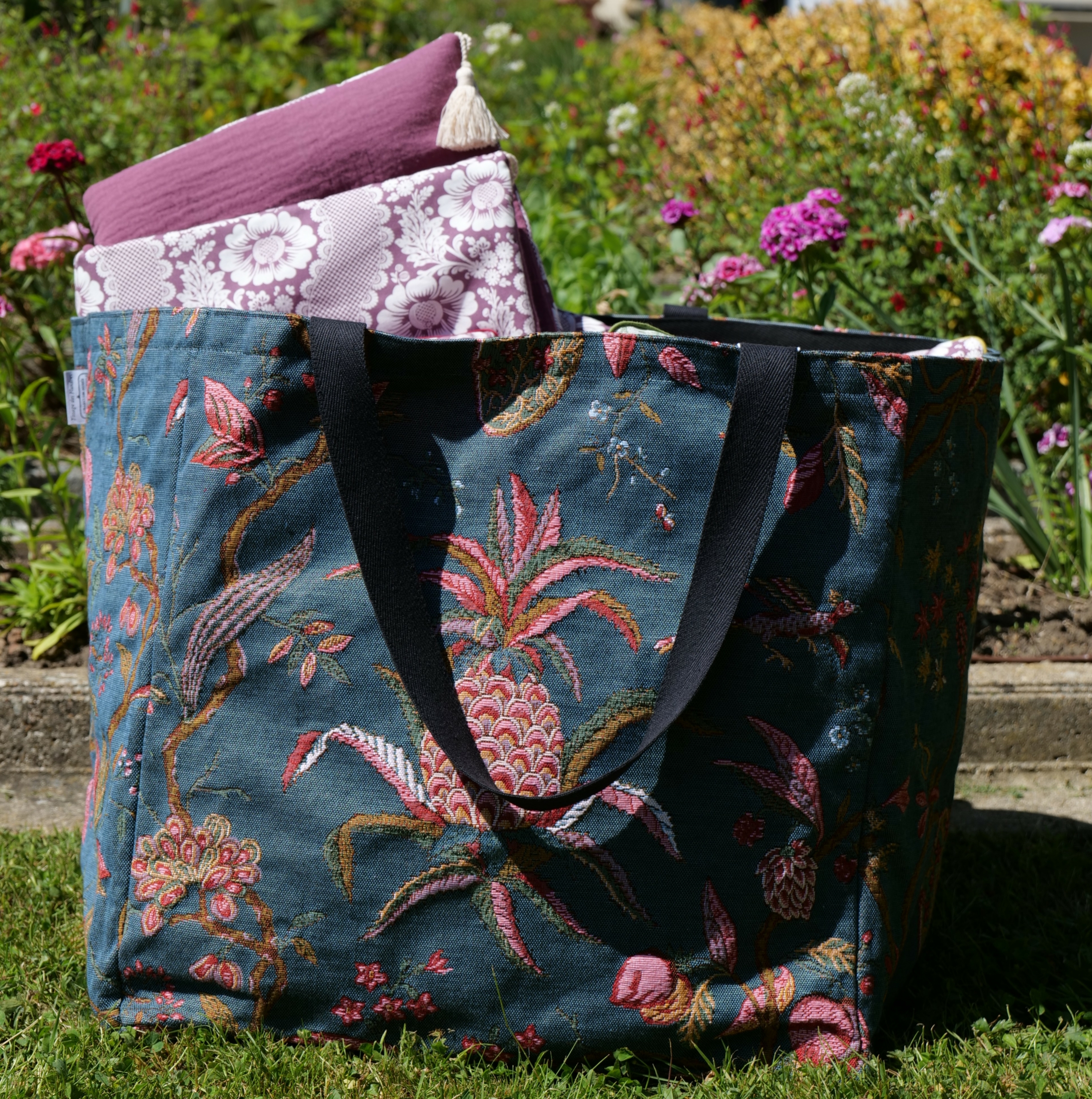 Le sac fourre-tout en tissu XCOMM_SAC CABAS GEANT ANANAS -TISSAGE est posé sur l'herbe d'un jardin ; il contient deux coussins à motifs violets et blancs, avec en arrière-plan des fleurs colorées et une végétation luxuriante. la marque Toile de Jouy