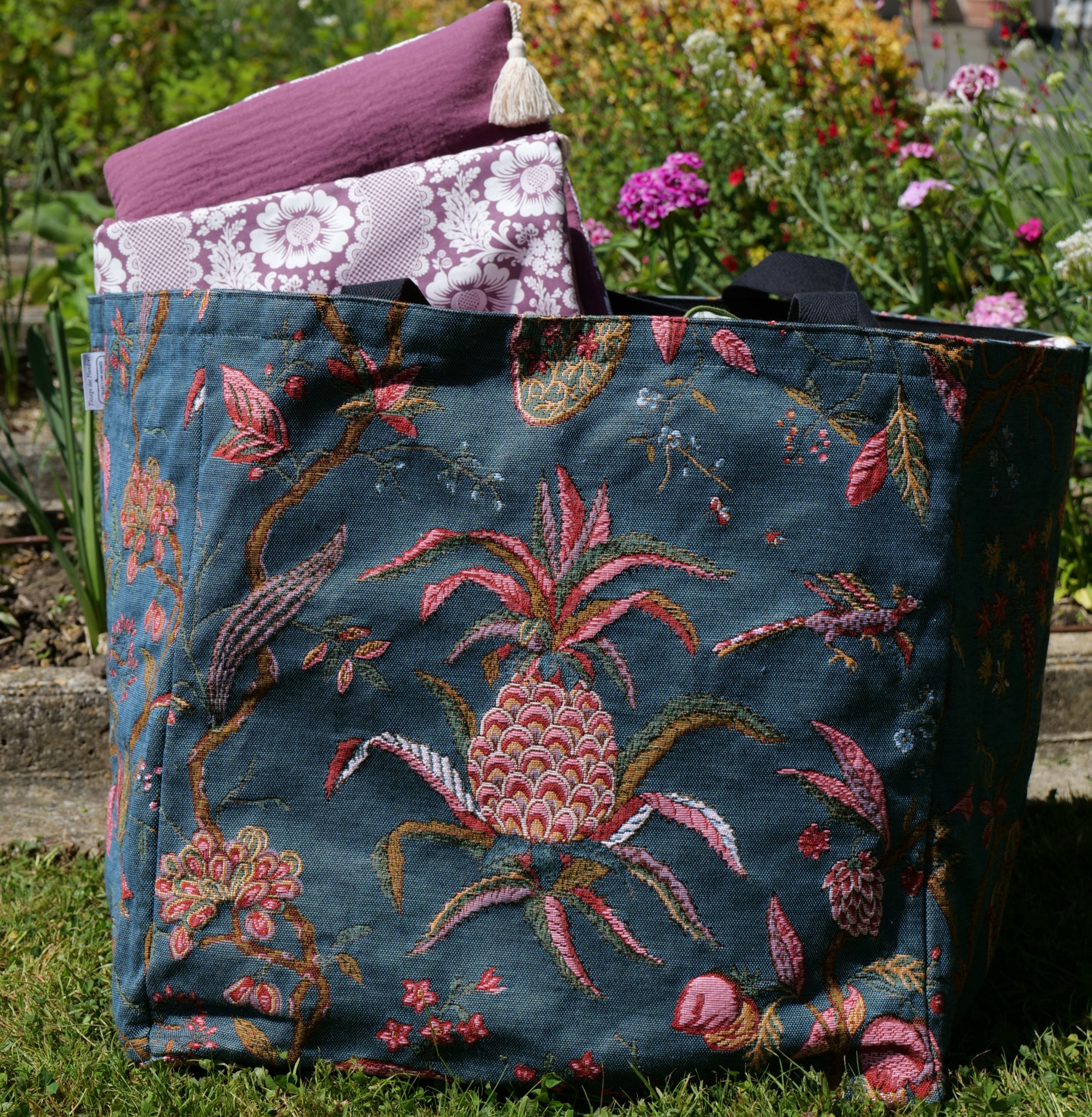 Le sac XCOMM_SAC CABAS GEANT ANANAS -TISSAGE, un grand sac fourre-tout bleu foncé à motifs floraux roses et verts, repose sur l'herbe d'un jardin, sur lequel sont posés deux coussins pliés aux motifs violets et blancs. Des fleurs colorées s'épanouissent en arrière-plan. la marque Toile de Jouy