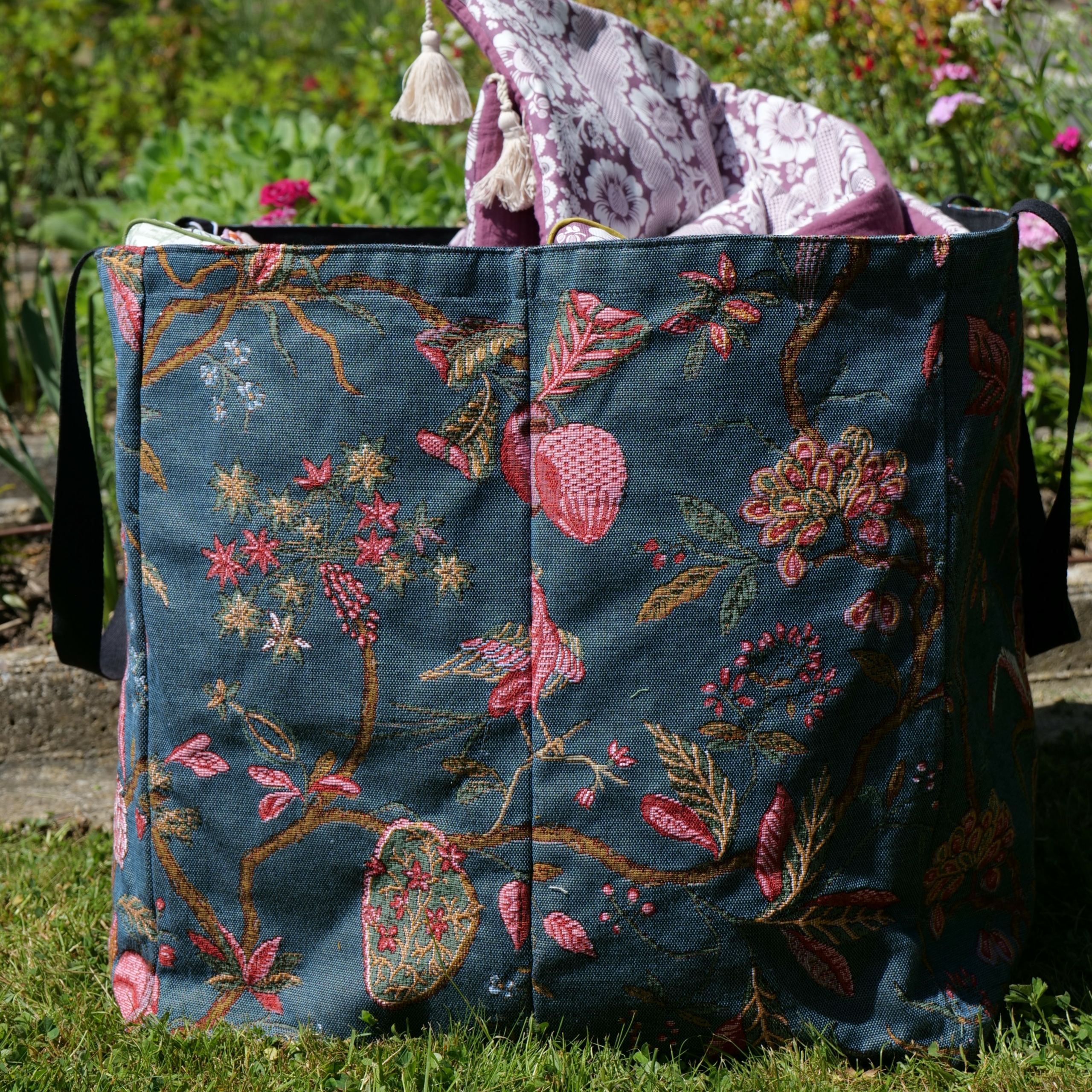 Le XCOMM_SAC CABAS GEANT ANANAS -TISSAGE, un grand sac fourre-tout en tissu orné de broderies florales et de papillons roses et verts, repose sur l'herbe, une couverture à motifs violets drapée sur son bord. On aperçoit des fleurs et de la verdure derrière lui. la marque Toile de Jouy