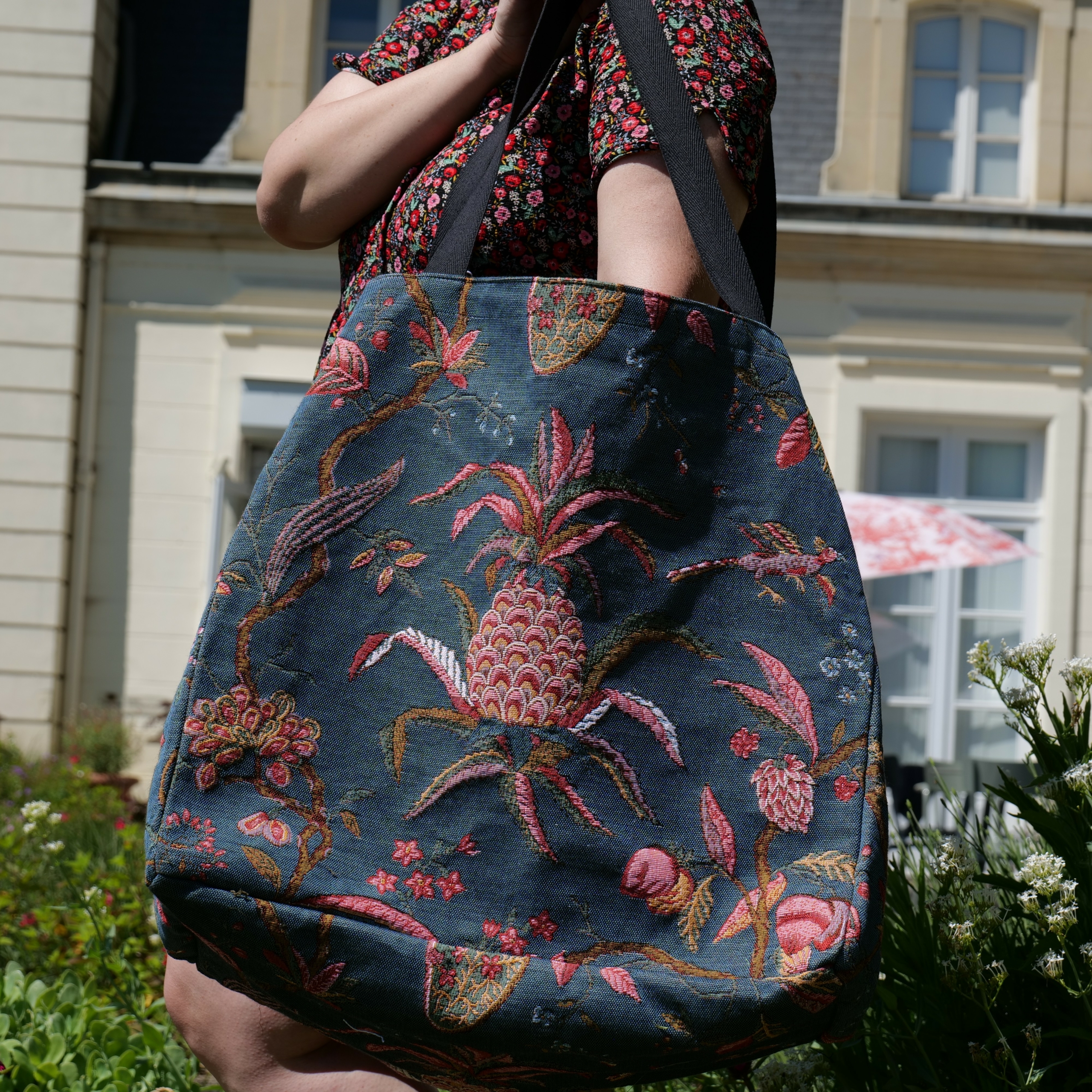 Une personne vêtue d'une robe à fleurs tient le XCOMM_SAC CABAS GEANT ANANAS -TISSAGE, un grand sac fourre-tout foncé orné de motifs botaniques et d'ananas brodés en rose et vert, debout à l'extérieur près d'un immeuble de couleur claire. la marque Toile de Jouy