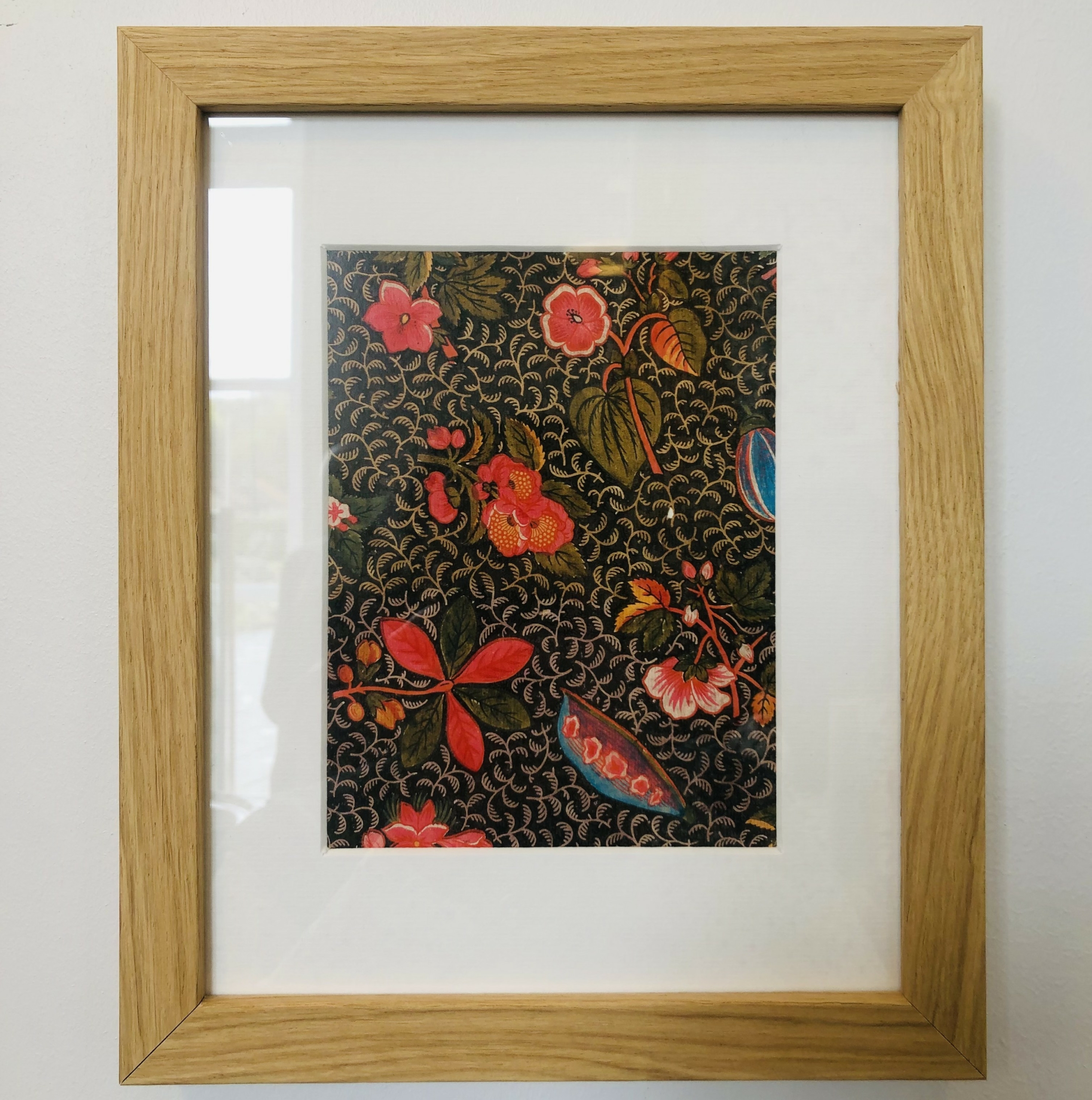 Le XCOMM_CARNET A5 BLEUETS présente des fleurs rouges, roses et orange vibrantes avec des feuilles vertes sur un fond sombre, présentées sous forme d'œuvre d'art encadrée avec un passe-partout blanc et un cadre en bois clair. la marque Toile de Jouy