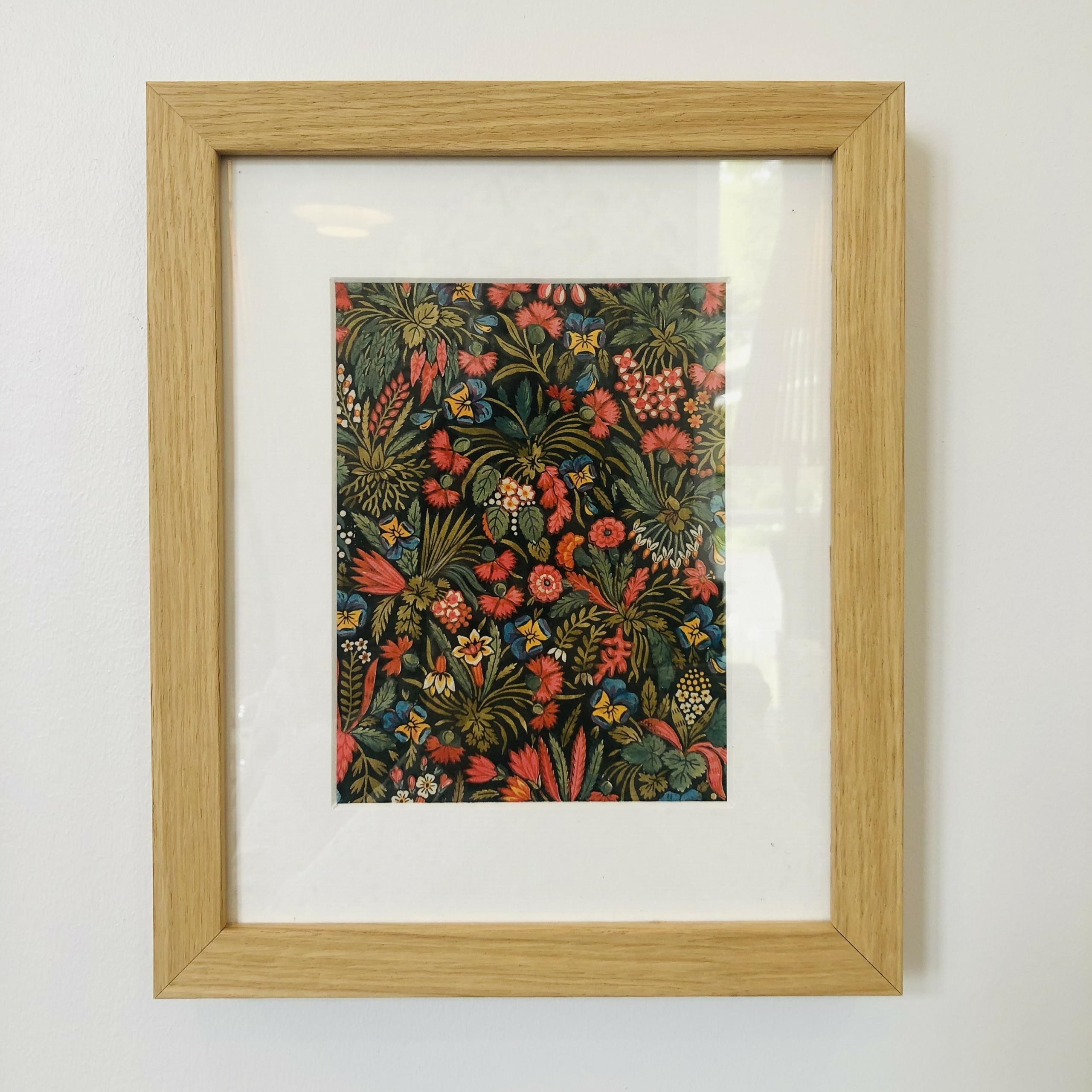 Le XCOMM_CARNET A5 BLEUETS présente un motif floral dense et vibrant avec des fleurs rouges, jaunes et vertes sur un fond sombre, magnifiquement encadré et exposé sur un mur blanc. la marque Toile de Jouy