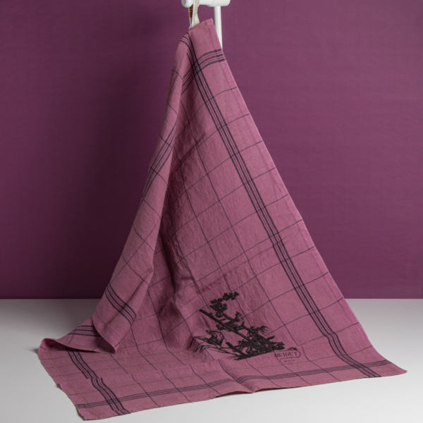 Le torchon XCOMM_TORCHON LES POULES MAUVE, qui présente un motif à carreaux violets avec des détails floraux noirs, est drapé sur un support blanc, sur un fond violet et une surface blanche assortis. la marque Toile de Jouy