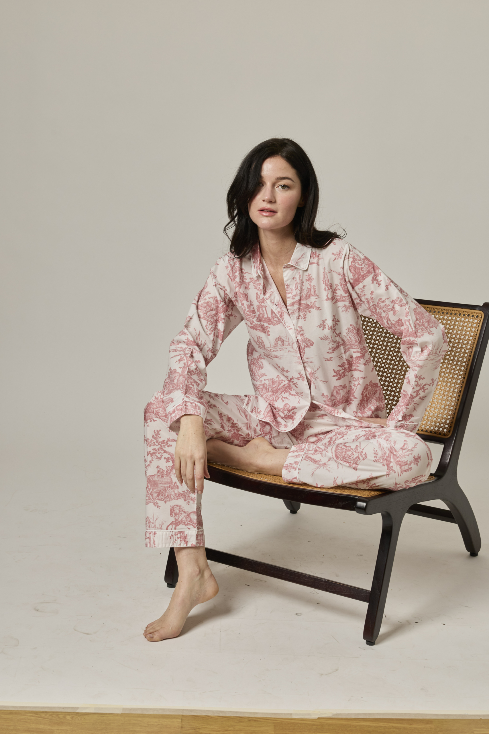 Une femme brune dans le PYJAMA COURT FEMME "LES DELICES DES 4 SAISONS" ROSE-ACANTHE est assise pieds nus sur une chaise en bois à dossier en forme de canne, regardant la caméra d'un air détendu dans un studio au décor minimal. la marque Toile de Jouy