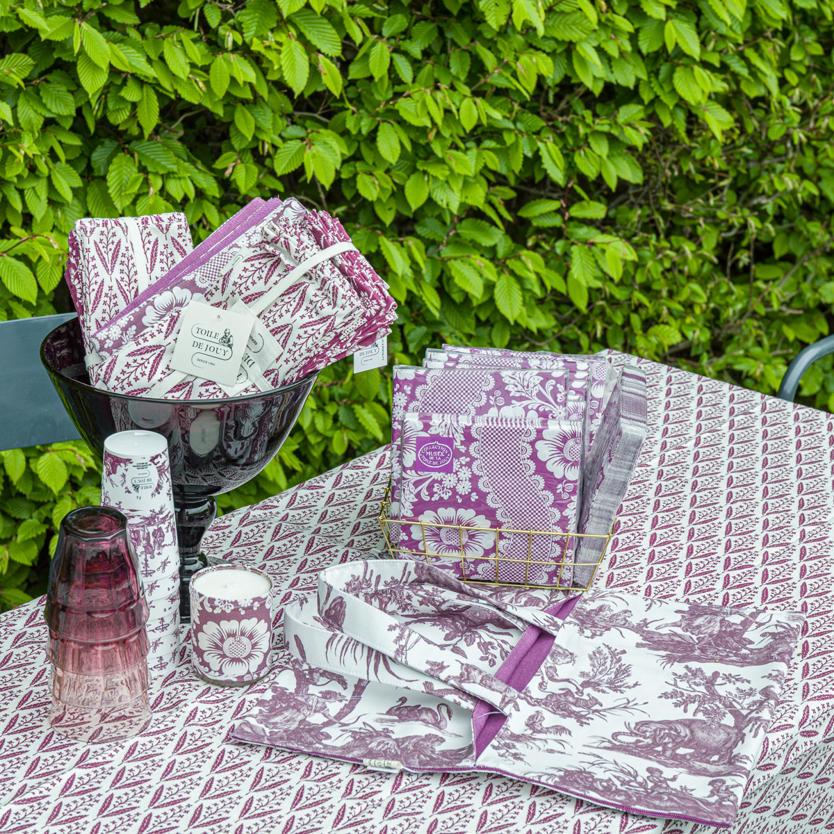 Le XCOMM_EMPORTE PLAT FLEURS AMARANTE est posé sur une table recouverte d'une nappe à motifs violets et blancs, avec des serviettes de table, des serviettes, des gobelets en papier et des verres assortis, le tout en plein air sur fond de haie verdoyante. la marque Toile de Jouy