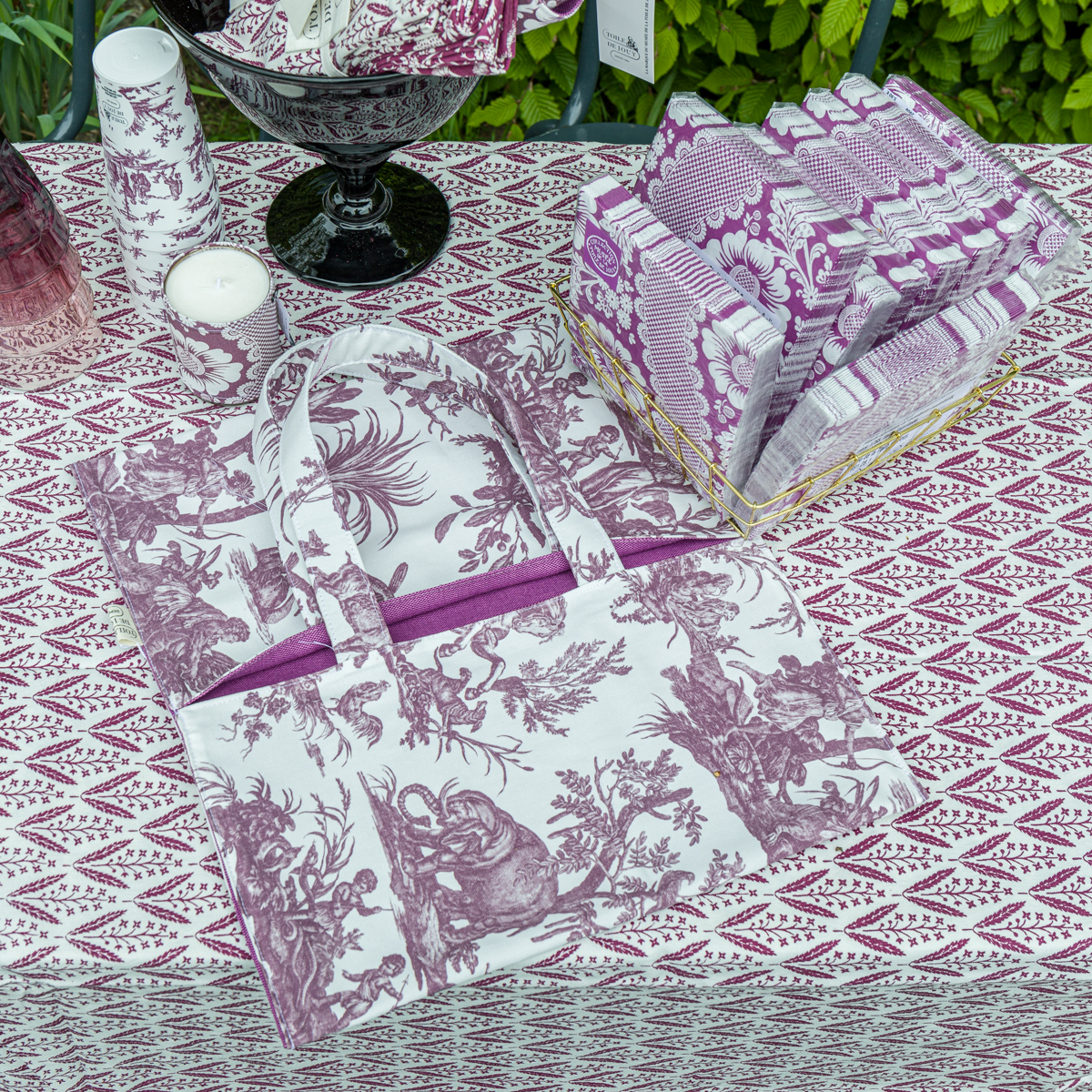 La nappe XCOMM_EMPORTE PLAT FLEURS AMARANTE ornée de motifs violets et blancs est présentée avec un sac en tissu assorti, des serviettes pliées, une bougie et un verre, sur fond de feuillage vert. la marque Toile de Jouy