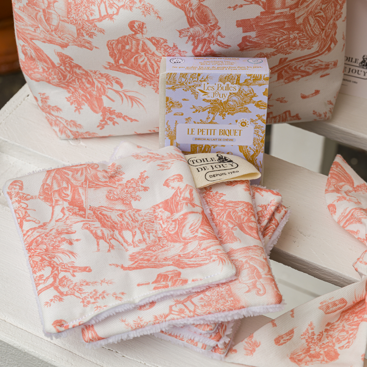 Un savon XCOMM_VANITY OFFRANDE SAUGE repose sur un tissu « toile » rose et blanc plissé, orné de scènes pastorales, posé sur une surface en bois blanc, avec une pochette à fermeture éclair assortie visible à l'arrière-plan. la marque Toile de Jouy