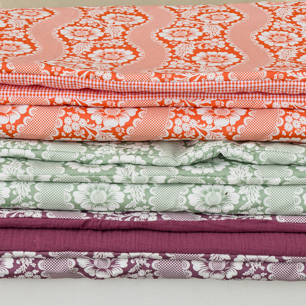 La collection XCOMM_MATELAS DE REPOS FLEURS STYLISEES MANDARINE 190X90 présente un ensemble de tissus orange, verts et violets soigneusement empilés, chacun orné de motifs floraux et géométriques blancs complexes sur un fond clair. la marque Toile de Jouy