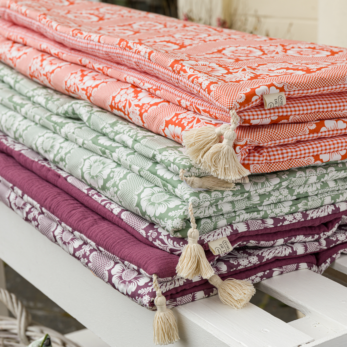 Le XCOMM_MATELAS DE REPOS FLEURS STYLISEES MANDARINE 190X90, avec un motif floral vibrant et des glands crème à une extrémité, est soigneusement plié et présenté sur une surface en bois blanc. la marque Toile de Jouy