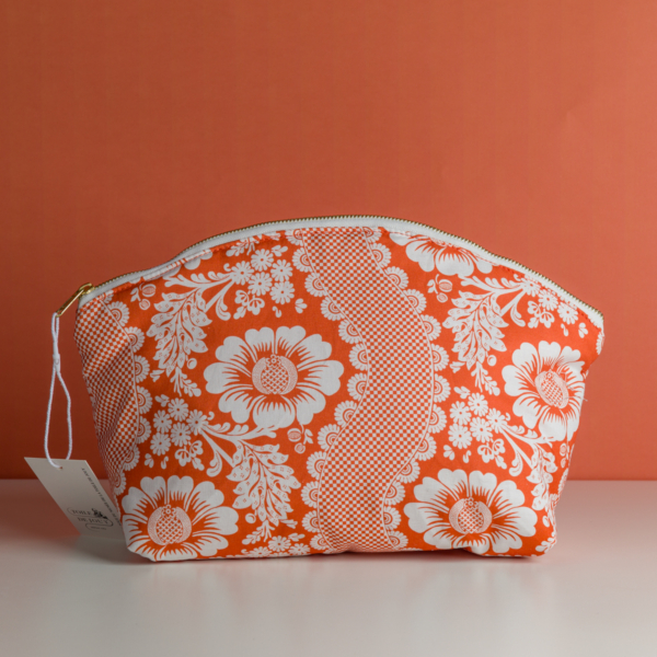 La pochette XCOMM_TROUSSE ARRONDIE FLEURS AMARANTE est une pochette à fermeture éclair ornée de motifs floraux blancs et de dentelle sur un fond orange vif. Elle est photographiée devant un mur orange, avec une étiquette blanche fixée à sa fermeture éclair. la marque Toile de Jouy