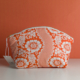 La pochette XCOMM_TROUSSE ARRONDIE FLEURS AMARANTE est une pochette à fermeture éclair ornée de motifs floraux blancs et de dentelle sur un fond orange vif. Elle est photographiée devant un mur orange, avec une étiquette blanche fixée à sa fermeture éclair. la marque Toile de Jouy