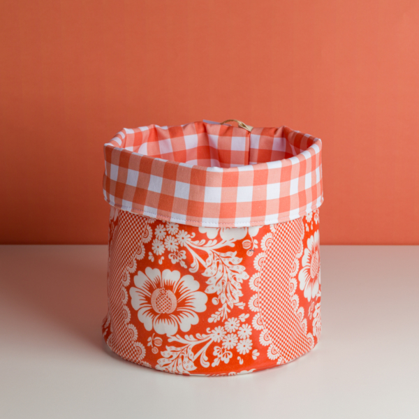 Le XCOMM_SET DE TABLE /2 FLEURS STYLISEES MANDARINE représente un panier en tissu à motifs floraux rouges et blancs avec un bord à carreaux vichy, sur une surface blanche à fond orange. la marque Toile de Jouy