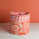 Le XCOMM_SET DE TABLE /2 FLEURS STYLISEES MANDARINE représente un panier en tissu à motifs floraux rouges et blancs avec un bord à carreaux vichy, sur une surface blanche à fond orange. la marque Toile de Jouy