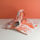 Le XCOMM_SET DE TABLE /2 FLEURS STYLISEES MANDARINE présente des motifs floraux et animaliers d'un orange vibrant, sur une surface blanche à fond pêche. Le design ouvert laisse entrevoir l'intérieur à motifs. la marque Toile de Jouy