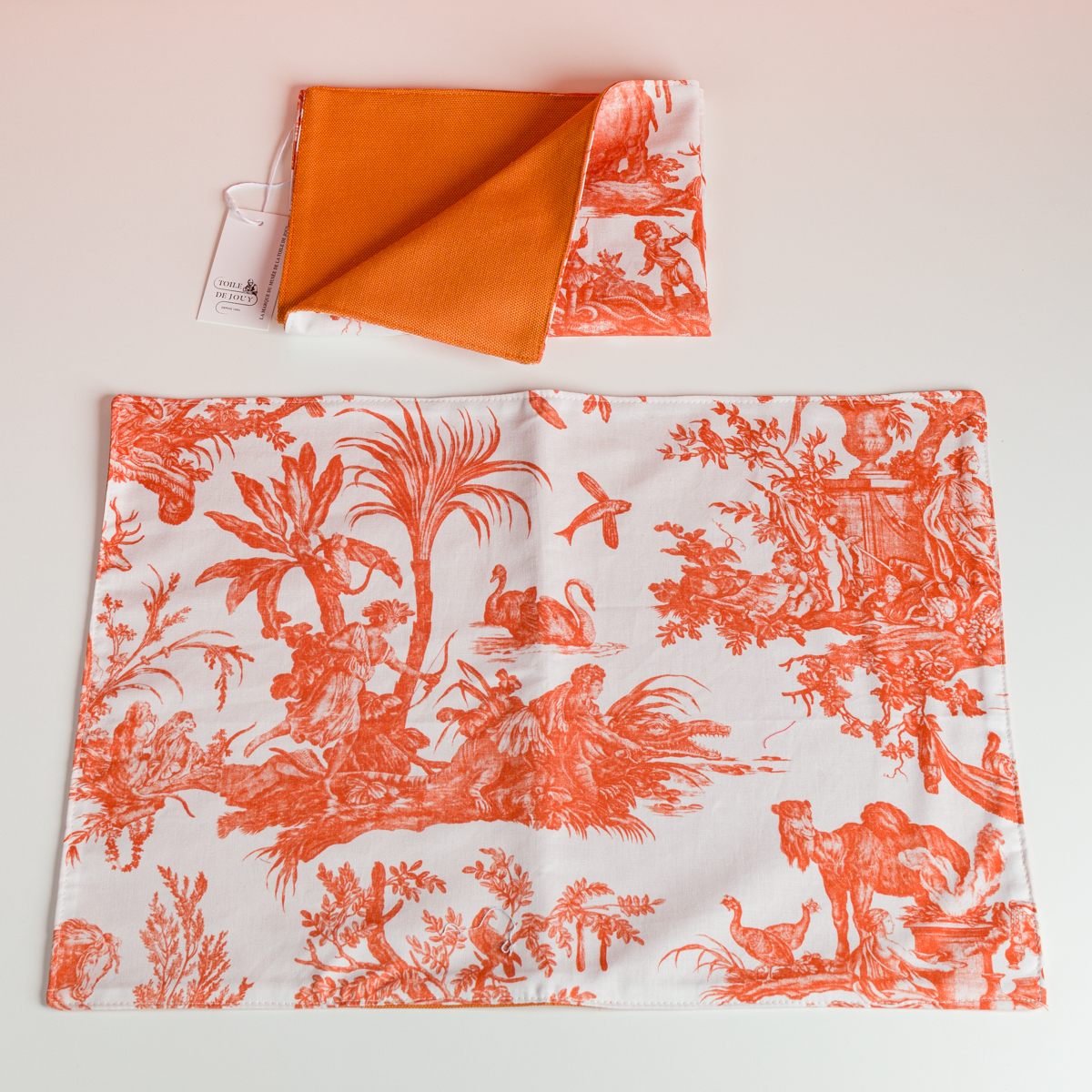 Le XCOMM_SET DE TABLE /2 FLEURS STYLISEES MANDARINE présente une serviette de table en tissu blanc avec un motif floral orange, étalée sur une surface, et une serviette pliée assortie au-dessus, montrant son verso en mandarine unie. la marque Toile de Jouy