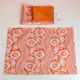 Le XCOMM_SET DE TABLE /2 FLEURS STYLISEES MANDARINE comprend un set de table floral orange et blanc aux bords festonnés présenté à plat, ainsi qu'une serviette de table pliée assortie aux côtés orange et fleuris avec une étiquette blanche attachée. la marque Toile de Jouy
