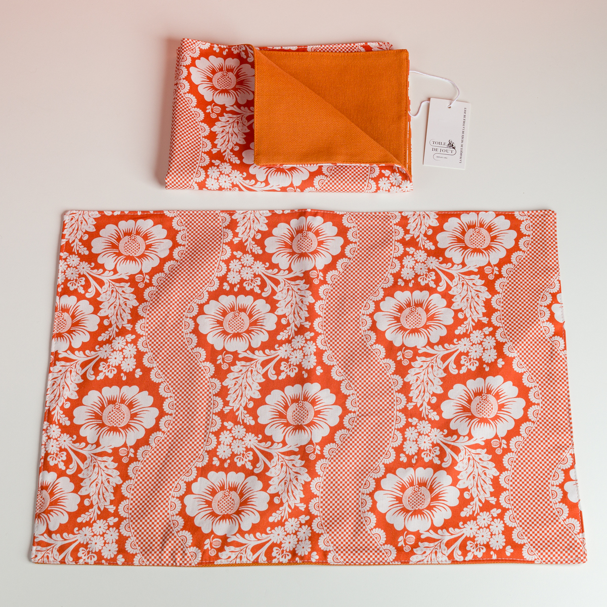 Le XCOMM_SET DE TABLE /2 FLEURS STYLISEES MANDARINE comprend un set de table floral orange et blanc aux bords festonnés présenté à plat, ainsi qu'une serviette de table pliée assortie aux côtés orange et fleuris avec une étiquette blanche attachée. la marque Toile de Jouy