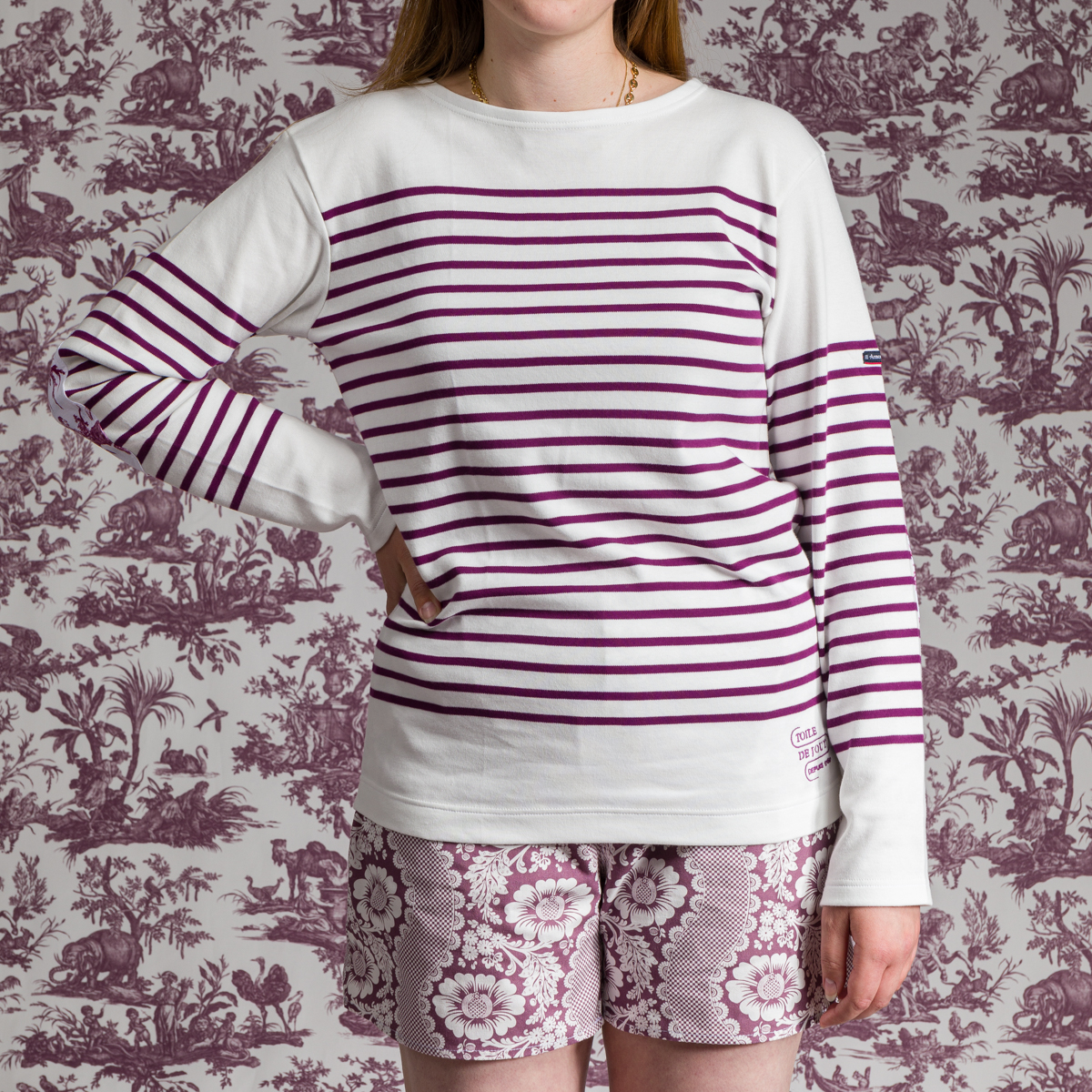 Une personne se tient devant un papier peint à motifs, portant la MARINIERE "4 PARTIES DU MONDE" ROSE PÊCHE - une chemise blanche à manches longues avec des rayures violettes et un short à fleurs violettes et blanches. Sa tête est partiellement hors du cadre. la marque Toile de Jouy