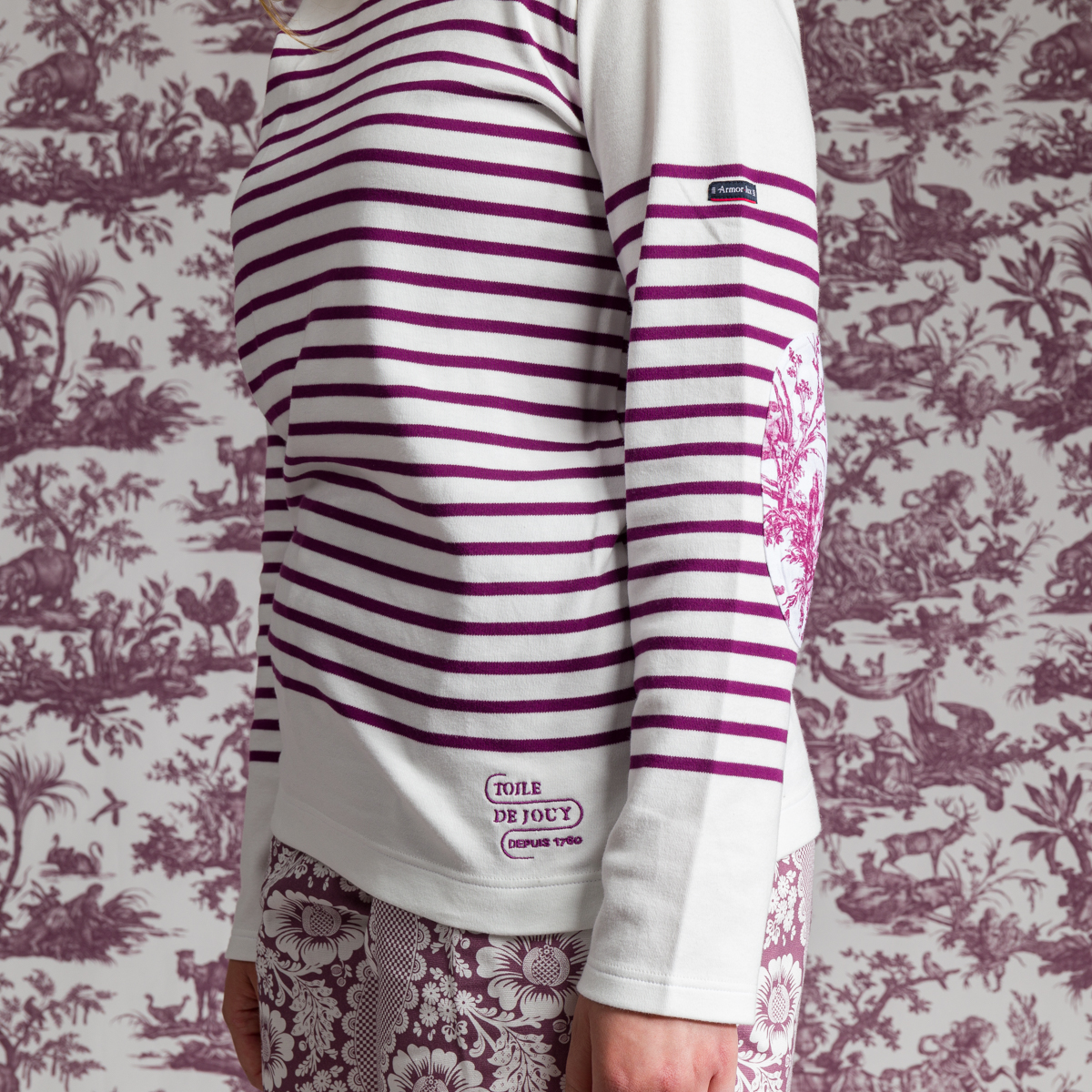Une personne modèle la MARINIERE "4 PARTIES DU MONDE" ROSE PÊCHE, une chemise blanche à manches longues avec des rayures et des motifs violets, devant un papier peint assorti de style toile, floral et pittoresque. Le visage de la personne n'est pas visible. la marque Toile de Jouy