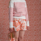 Une femme se tient pieds nus sur un fond rose et blanc, vêtue de la chemise à manches longues rayée MARINIERE "4 PARTIES DU MONDE" ROSE PÊCHE, d'un short imprimé et de chaussures à enfiler assorties, tous dotés de motifs coordonnés. la marque Toile de Jouy