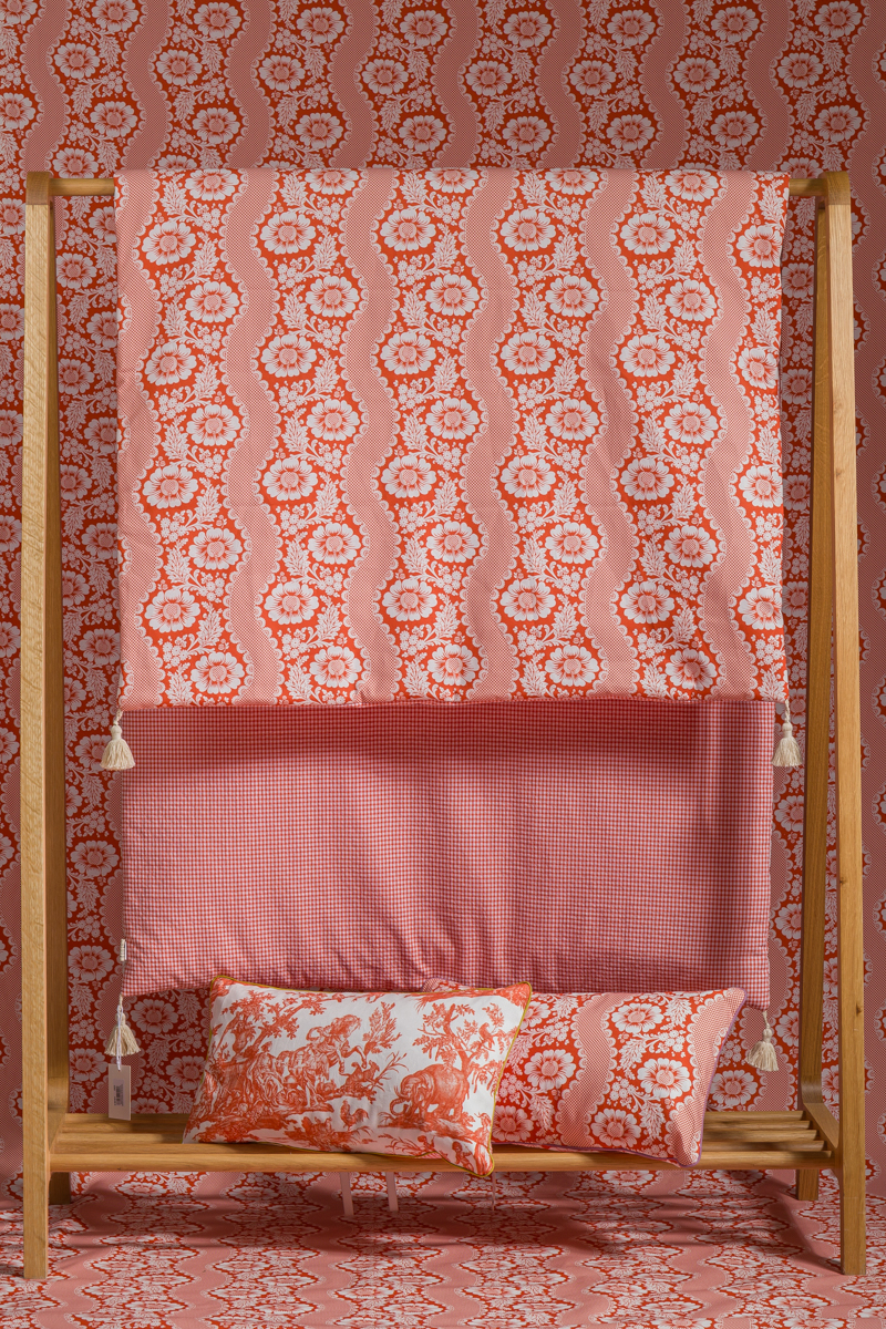 Une étagère en bois présente le tapis XCOMM_MATELAS DE REPOS FLEURS STYLISEES MANDARINE 190X90, un tapis à motifs roses et rouges avec une décoration florale ondulée, ainsi que des textiles coordonnés et un papier peint floral assorti à l'arrière-plan. la marque Toile de Jouy