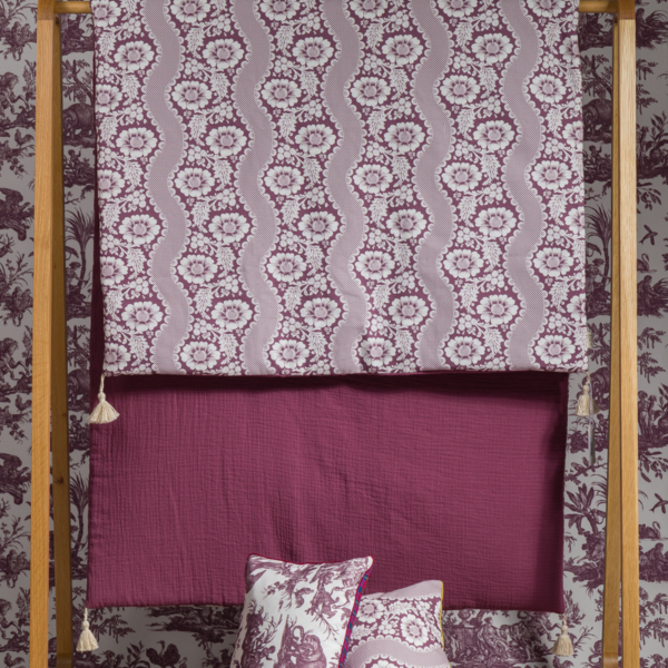 Le XCOMM_MATELAS DE REPOS FLEURS STYLISEES AMARANTE 190X90 est posé sur un support en bois à côté de couvertures bordeaux, avec des oreillers bordeaux et blancs assortis devant, le tout sur fond de papier peint à motifs. la marque Toile de Jouy