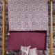 Le XCOMM_MATELAS DE REPOS FLEURS STYLISEES AMARANTE 190X90 est posé sur un support en bois à côté de couvertures bordeaux, avec des oreillers bordeaux et blancs assortis devant, le tout sur fond de papier peint à motifs. la marque Toile de Jouy