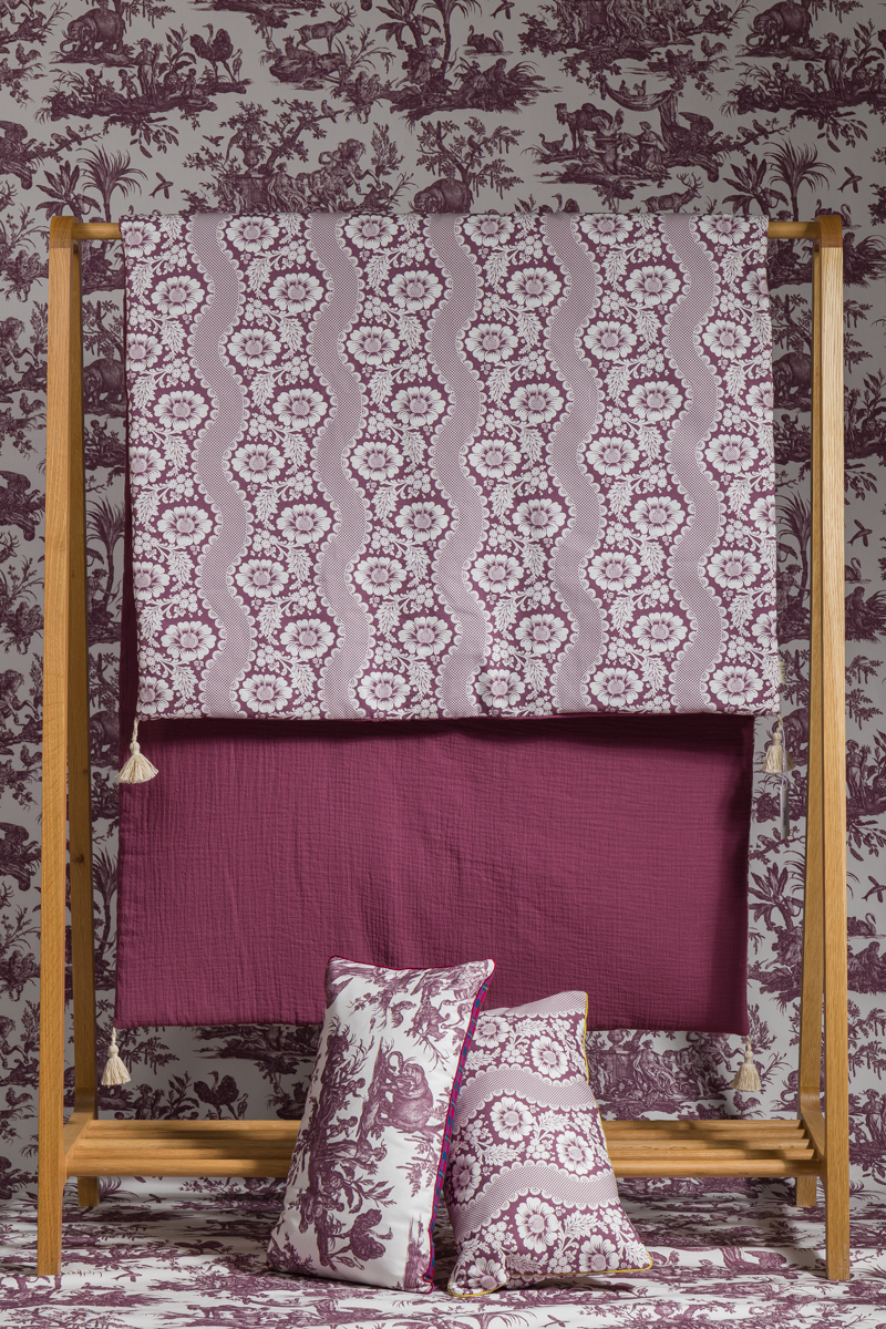 Le XCOMM_MATELAS DE REPOS FLEURS STYLISEES AMARANTE 190X90 est posé sur un support en bois à côté de couvertures bordeaux, avec des oreillers bordeaux et blancs assortis devant, le tout sur fond de papier peint à motifs. la marque Toile de Jouy