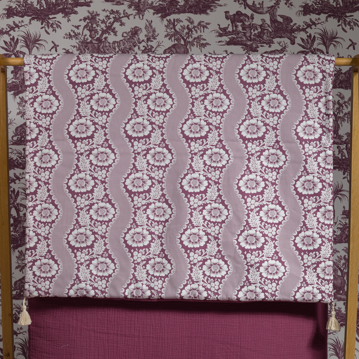 Le XCOMM_MATELAS DE REPOS FLEURS STYLISEES AMARANTE 190X90, avec un motif floral stylisé violet et blanc, est drapé sur un cadre en bois. Le papier peint Toile aux motifs champêtres violets assortis forme l'arrière-plan. la marque Toile de Jouy