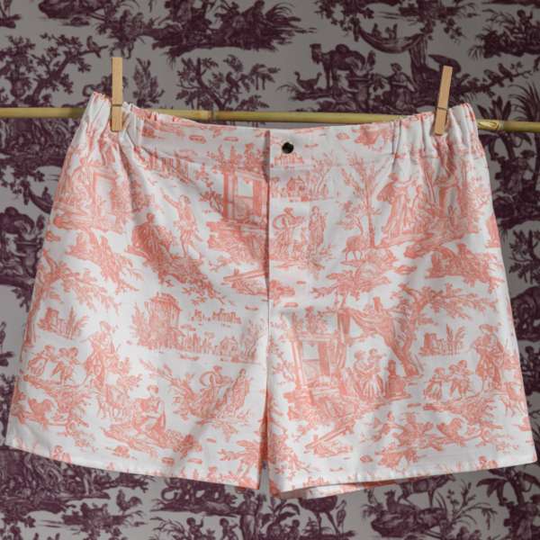 Le short CALEçON « FLEURS STYLISÉES » SAUGE, sur fond blanc orné d’un imprimé « toile » champêtre rose, est suspendu à des pinces à linge fixées sur une tringle en bois. L’arrière-plan présente un motif « toile » violet foncé assorti. la marque Toile de Jouy