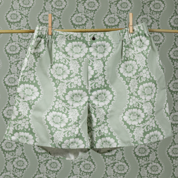 Le short CALEçON « FLEURS STYLISÉES » SAUGE, vert et blanc, est suspendu à une corde à linge devant un mur recouvert d'un papier peint à motifs floraux assortis, les deux éléments se fondant l'un dans l'autre pour créer un effet de camouflage. la marque Toile de Jouy
