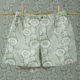 Le short CALEçON « FLEURS STYLISÉES » SAUGE, vert et blanc, est suspendu à une corde à linge devant un mur recouvert d'un papier peint à motifs floraux assortis, les deux éléments se fondant l'un dans l'autre pour créer un effet de camouflage. la marque Toile de Jouy